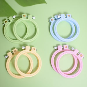 May include: Four sets of embroidery hoops in pastel colors: green, blue, yellow, and pink. Each set contains two hoops, with a metal screw and tightening mechanism. The hoops are arranged on a light green surface, with a green leaf in the background.