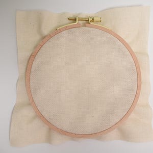 May include: A round embroidery hoop with a beige fabric stretched tightly across the frame. The hoop is made of natural wood and has a brass screw to tighten the fabric.
