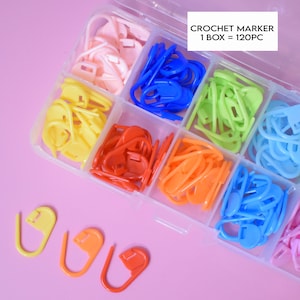 May include: A clear plastic box filled with 120 colorful plastic crochet stitch markers. The box has 8 compartments, each filled with a different color of markers. The colors are pink, blue, green, yellow, red, orange, light blue, and pink. The box has a label that reads "CROCHET MARKER 1 BOX = 120PC".