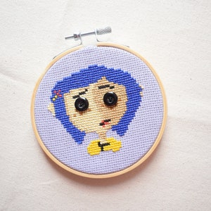 May include: A cross-stitch embroidery hoop with a purple background and a cartoon image of a girl with blue hair, yellow shirt, and black button eyes.