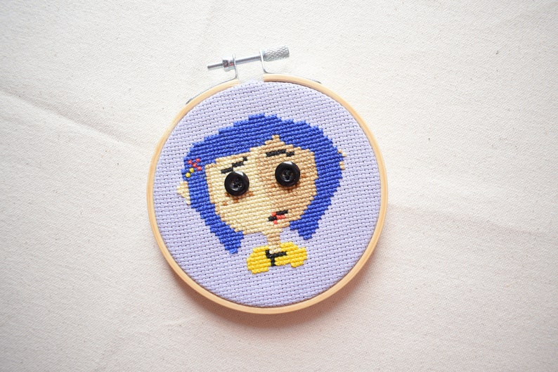 May include: A cross-stitch embroidery hoop with a purple background and a cartoon image of a girl with blue hair, yellow shirt, and black button eyes.