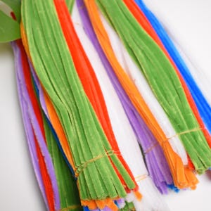 May include: A collection of colorful craft supplies, including green, orange, red, white, purple, and blue pipe cleaners. The pipe cleaners are bundled together with twine and arranged against a white background. A green leaf is in the upper left corner.