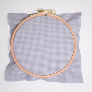 Può includere: Un telaio da ricamo di colore grigio chiaro con una cornice in legno e un tessuto grigio chiaro teso sopra.