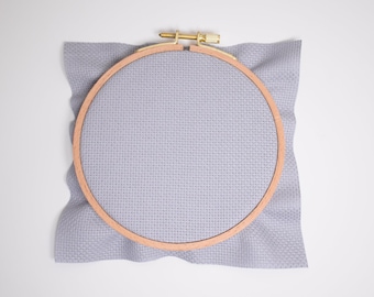 May include: A light grey embroidery hoop with a wooden frame and a light grey fabric stretched across it.