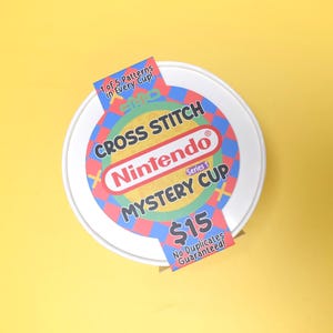 May include: A white, round container with a colorful label featuring the Nintendo logo and the words "Cross Stitch Mystery Cup." The label includes text that reads "1 of 5 Patterns in Every Cup!" and "No Duplicates Guaranteed!"