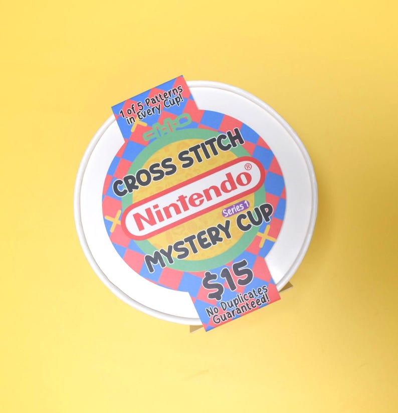 May include: A white, round container with a colorful label featuring the Nintendo logo and the words "Cross Stitch Mystery Cup." The label includes text that reads "1 of 5 Patterns in Every Cup!" and "No Duplicates Guaranteed!"