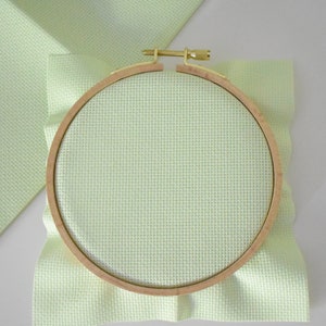 May include: A wooden embroidery hoop holds light green cross-stitch fabric, with a matching piece of fabric in the background. The hoop is secured with a brass screw. The fabric has a grid-like pattern, ideal for needlework projects.