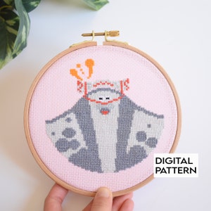 May include: A cross-stitch pattern of a gray manta ray with orange spots on a pink background. The manta ray is wearing a red and white hat with orange spots. The pattern is framed in a wooden embroidery hoop.