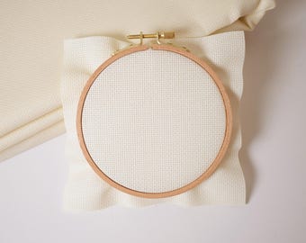 May include: A wooden embroidery hoop with a brass clasp holds a piece of cream-coloured cross-stitch fabric. The fabric is stretched taut within the hoop, ready for stitching. Additional folded fabric is visible in the background, suggesting a crafting project.