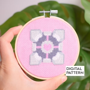 Könnte beinhalten: Ein pinkfarbenes und graues Kreuzstichmuster eines Portals aus dem Videospiel Portal. Das Muster ist in einem Holzrahmen eingefasst. Der Text "DIGITAL PATTERN" ist unter dem Rahmen gedruckt.
