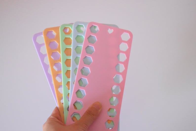 K&ouml;nnte beinhalten: Eine Hand h&auml;lt einen F&auml;cher aus farbigen Kunststoff-Fadenorganisatoren. Die Organisatoren sind in den Farbt&ouml;nen Lavendel, Orange, Gr&uuml;n und Rosa gehalten. Jeder Organizer hat eine Reihe sechseckiger Aussparungen und ein herzf&ouml;rmiges Loch oben.