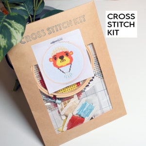 May include: A cross stitch kit featuring a cartoon animal with a red and yellow face, wearing a yellow hat and a blue necklace. The kit includes a hoop, fabric, and embroidery floss.
