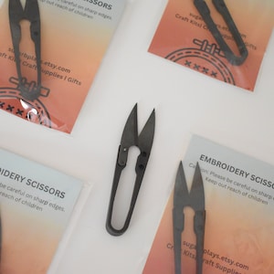 Puede incluir: Cuatro pares de tijeras de bordado negras con una etiqueta blanca que dice "Tijeras de bordado" y "Precaución: Tenga cuidado con los bordes afilados. Mantenga fuera del alcance de los niños". La etiqueta tiene un fondo melocotón con un degradado rojo y naranja.