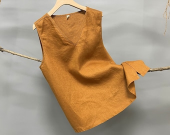 May include: A sleeveless, V-neck top made of linen in a burnt orange color. The top is hanging on a wooden branch.