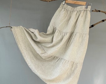 Op de afbeelding: Een beige linnen rok met tierlagen en een geplooide tailleband. De rok hangt aan een houten hanger en waait in de lucht.