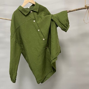 May include: Olive green button-down shirt with long sleeves, hanging on a wooden hanger. The shirt has a relaxed fit and a unique asymmetrical design. The fabric appears to be linen or a similar lightweight material.