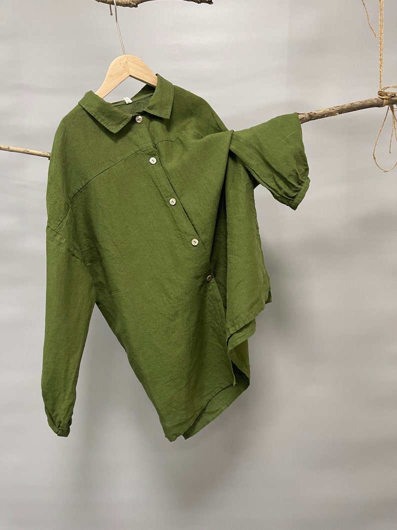 May include: Olive green button-down shirt with long sleeves, hanging on a wooden hanger. The shirt has a relaxed fit and a unique asymmetrical design. The fabric appears to be linen or a similar lightweight material.