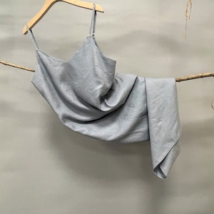 May include: A light gray-blue slip dress with thin straps hangs from a wooden hanger. The dress is made of a soft, flowing fabric and is draped over a branch. The color is described as "Grayish Blue" in the image.