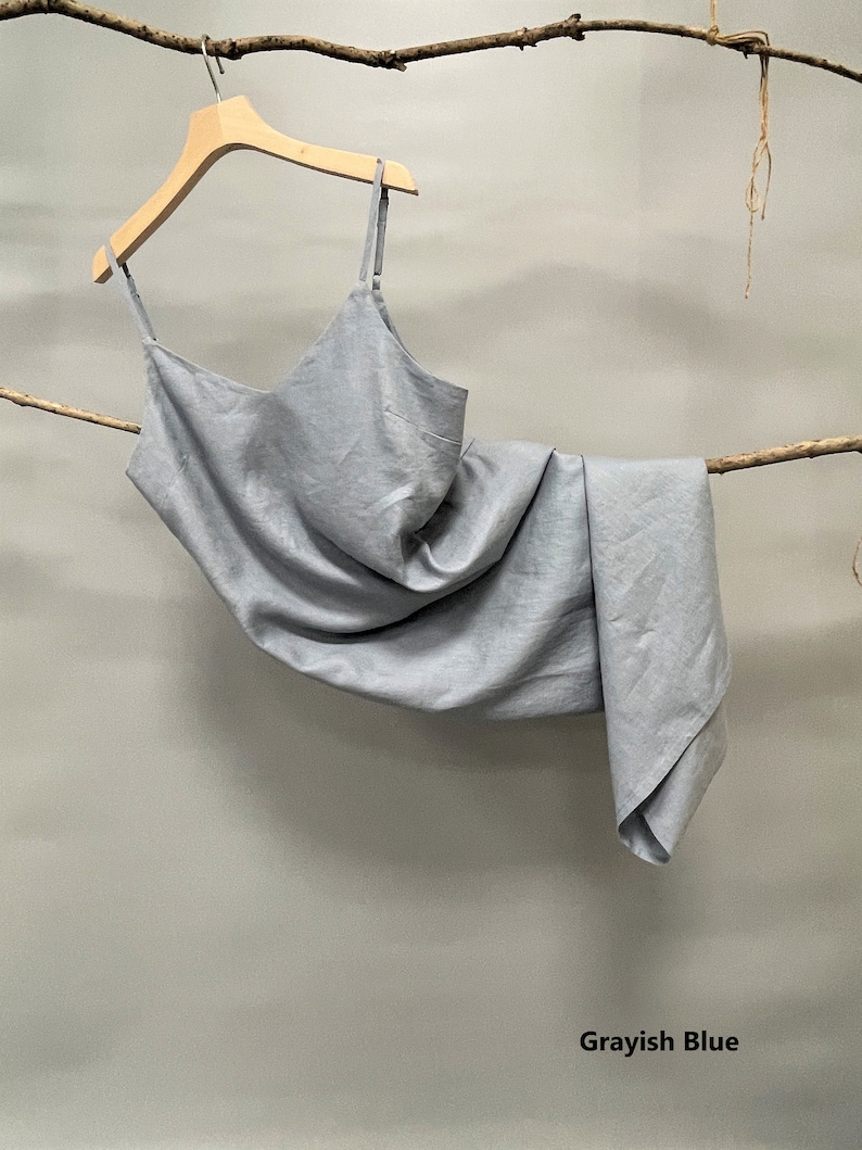 May include: A light gray-blue slip dress with thin straps hangs from a wooden hanger. The dress is made of a soft, flowing fabric and is draped over a branch. The color is described as "Grayish Blue" in the image.