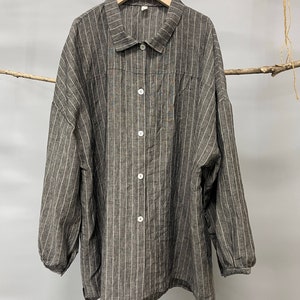 May include: Gray pinstripe button-down shirt with long sleeves. The shirt features vertical white stripes on a gray background. It has a collared neckline and white buttons. The shirt is hanging on a wooden hanger.