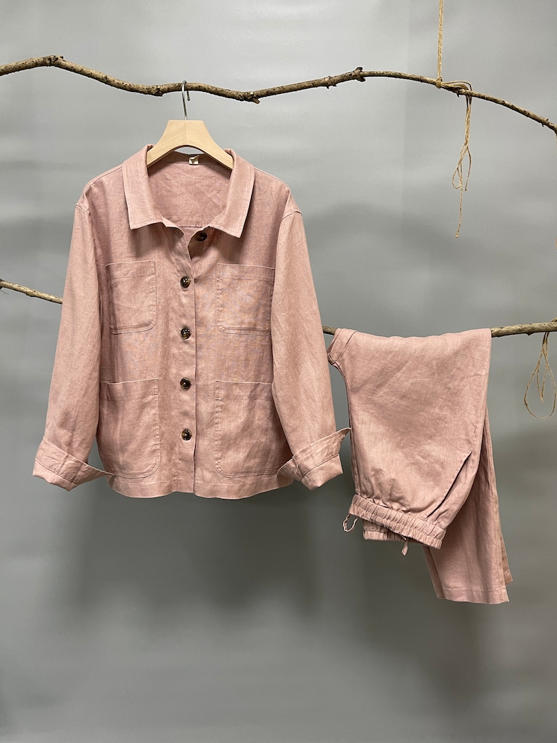 Puede incluir: Conjunto de dos piezas de lino rosa claro, que incluye una chaqueta abotonada con dos bolsillos delanteros y pantalones a juego con cintura con cord&oacute;n. La chaqueta tiene mangas largas y botones de madera. El conjunto se exhibe en una percha de madera.