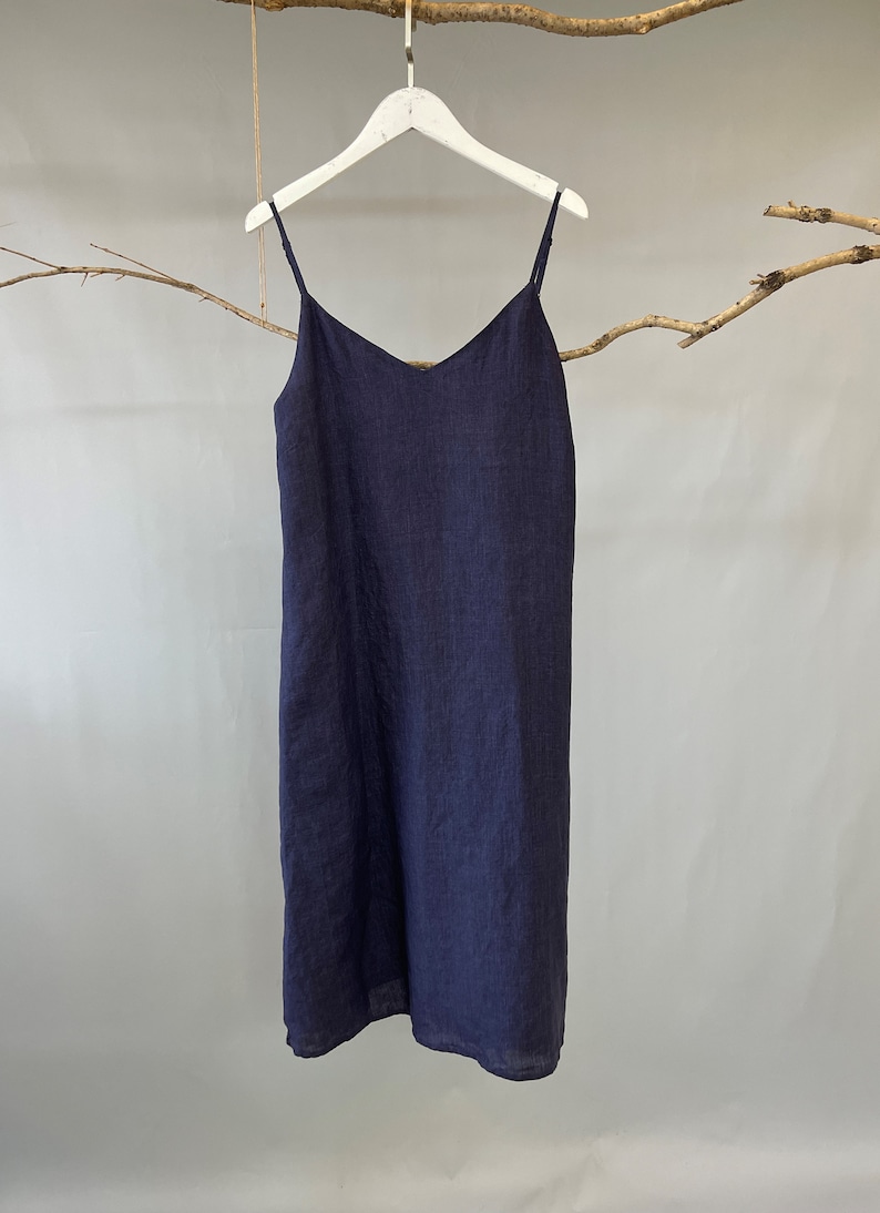 May include: A navy blue slip dress with thin spaghetti straps and a V-neck. The dress is made of a lightweight fabric and hangs on a white hanger. The dress is approximately knee-length.
