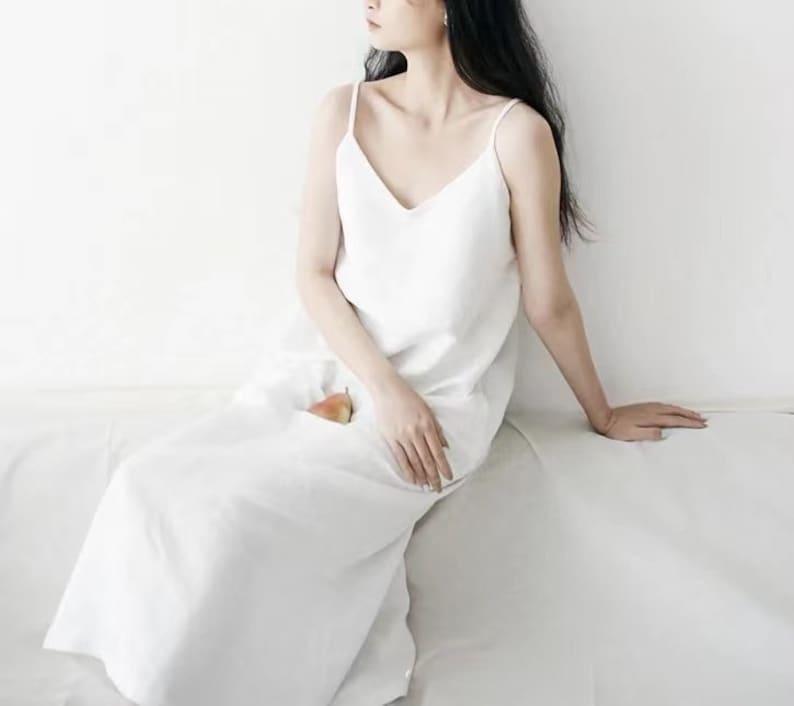 May include: A white, V-neck slip dress with thin straps. The dress is long and flowing, and a pear is tucked into a pocket. The model is sitting on a white surface.