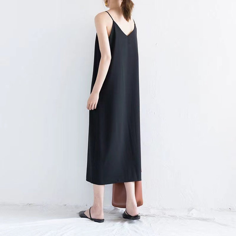 May include: A black, sleeveless slip dress with a V-neck. The dress is ankle-length and has thin straps. The person is wearing black sandals and carrying a brown tote bag. The background is a plain white wall.