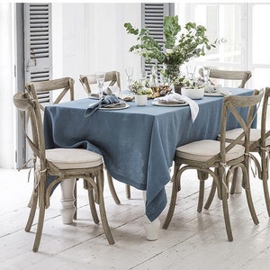 May include: A dining table set for a meal, featuring a blue linen tablecloth and six wooden chairs with cream-colored cushions. The table is set with plates, napkins, and glassware, and a vase of greenery sits in the center. The chairs have a cross-back design.