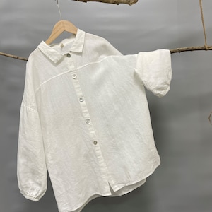 May include: A white linen button-down shirt with long sleeves and a classic collar. The shirt is hanging on a wooden hanger, with a natural branch in the background. The shirt has a relaxed fit and a slightly textured appearance.