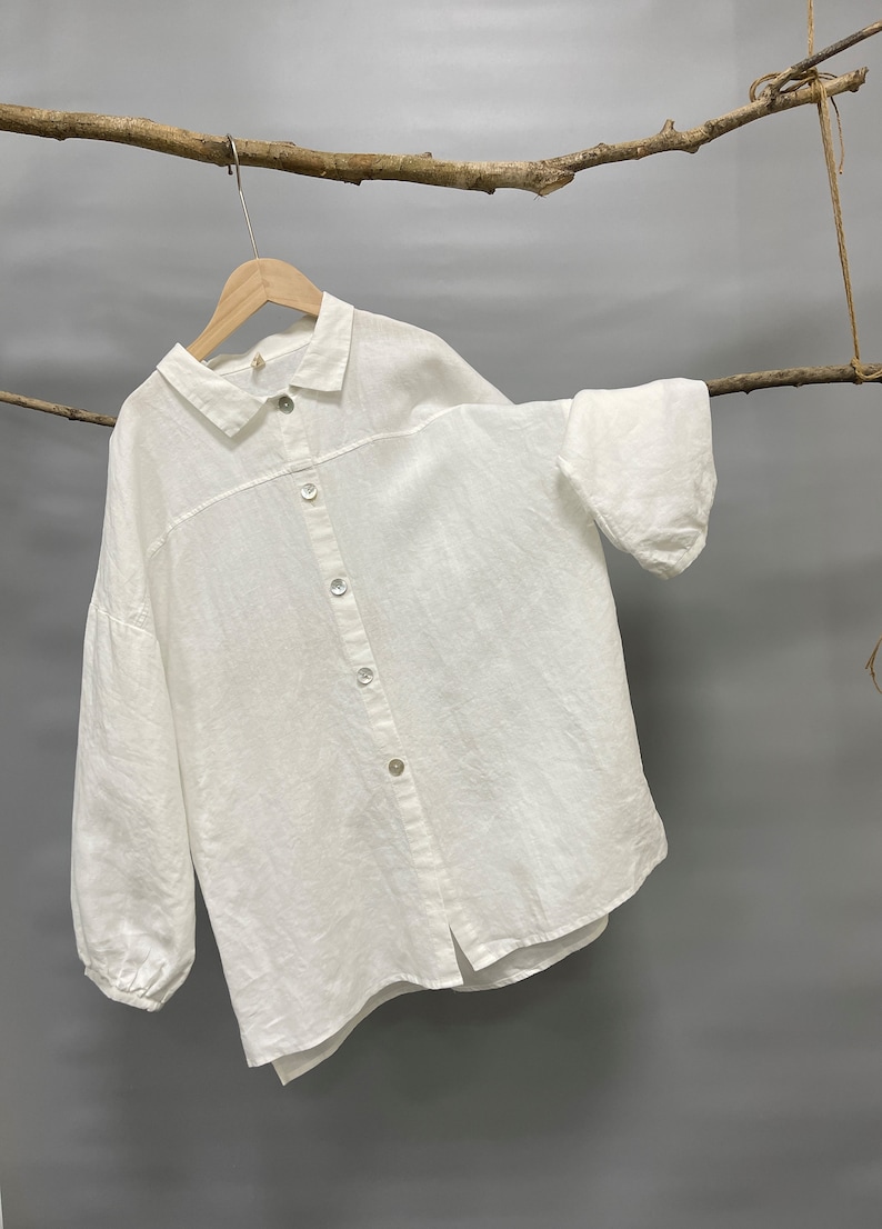 May include: A white linen button-down shirt with long sleeves and a classic collar. The shirt is hanging on a wooden hanger, with a natural branch in the background. The shirt has a relaxed fit and a slightly textured appearance.