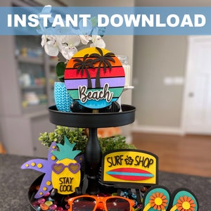 May include: A tiered tray with a beach theme. The top tier has a round wooden sign with a sunset and palm trees, and the text "Beach". The middle tier has a pineapple with sunglasses and the text "Stay Cool". The bottom tier has a surfboard with the text "Surf Shop", a pair of flip flops, and sunglasses.