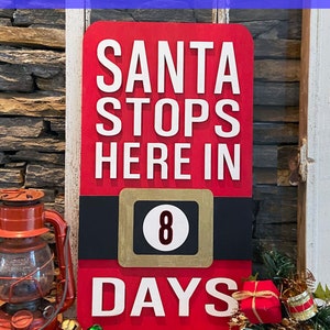May include: Red wooden sign with white text that reads "Santa Stops Here In 8 Days". The sign has a black belt with a gold buckle and a gold circle with the number 8 inside.