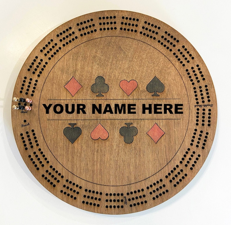 May include: A wooden cribbage board with a brown stain. The board has a circular shape with a black border and black dots around the edge. The center of the board has a white area with the text "YOUR NAME HERE" and four playing card symbols. The symbols are a red diamond, a black club, a red heart, and a black spade.