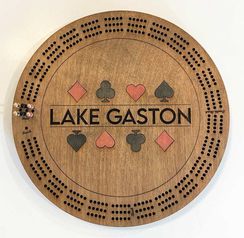 May include: A wooden cribbage board with a brown stain. The board has a circular shape with a black border and black dots around the edge. The center of the board has the text "LAKE GASTON" in black lettering. There are four red and four black playing card symbols in the center of the board.