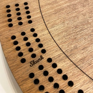 May include: A wooden circular board with a pattern of holes around the edge. The board is engraved with the word "Skunk".