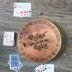 May include: A wooden cribbage board with the text "Relax you're at the Lake" on it. There are playing cards scattered around the board.