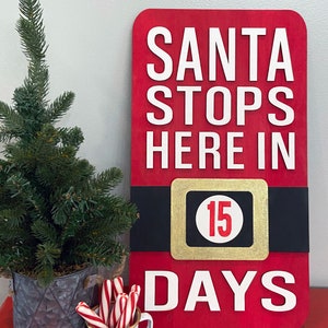 May include: A red wooden sign with white text that reads "Santa Stops Here In 15 Days". The sign is decorated with a black and gold belt with a white circle containing the number 15. The sign is sitting on a red table with a small potted tree and a mug with candy canes.