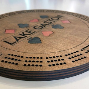 May include: A wooden cribbage board with a brown stain. The board has a circular shape and features a personalized inscription "LAKE GASTON" with four playing card suits in the center. The board has a series of holes around the edge for playing cribbage.