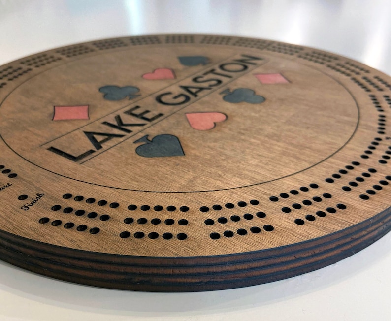 May include: A wooden cribbage board with a brown stain. The board has a circular shape and features a personalized inscription "LAKE GASTON" with four playing card suits in the center. The board has a series of holes around the edge for playing cribbage.