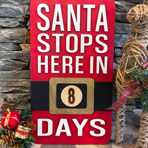 May include: Red wooden sign with the text "SANTA STOPS HERE IN" and a number "8" in a gold circle. The sign also says "DAYS" in white letters. The sign is leaning against a stone wall with a brown and red plaid ribbon and a brown rope reindeer decoration.