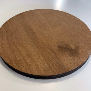 May include: A round wooden coaster with a brown woodgrain finish. The coaster is made of multiple layers of wood and has a slightly raised edge.