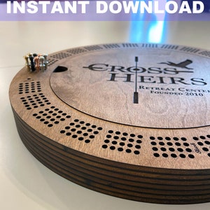 May include: A wooden game board with a circular design and a center section that lifts up. The board is engraved with the words "Cross Heirs Retreat Center Founded 2010".