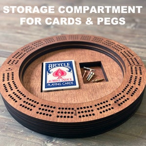 May include: A wooden cribbage board with a storage compartment for cards and pegs. The board is made of dark wood and has a circular shape. The compartment is located on the side of the board and is large enough to hold a deck of cards and a set of pegs. The board is perfect for playing cribbage with friends and family.