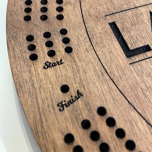 May include: A wooden game board with a circular shape and a brown stain. The board has a series of holes in a circular pattern around the edge. The words "Start" and "Finish" are engraved on the board. The board is made of wood and has a natural grain.