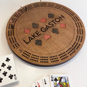May include: A wooden cribbage board with a brown stain. The board has a circular shape with a center circle and multiple rows of holes around the edge. The board is personalized with the text "LAKE GASTON" and four playing card suits.