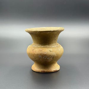 Puede incluir: Un pequeño jarrón de estilo antiguo con una superficie texturizada de color marrón claro. El jarrón tiene una base redondeada, una sección media bulbosa y un borde ensanchado. La forma general es simétrica y el jarrón parece estar hecho de arcilla o un material similar.