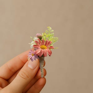 May include: A small, handcrafted boutonniere featuring a cluster of colorful flowers. The arrangement includes a large, coral-colored daisy with a yellow center, a smaller purple flower, and delicate green foliage, all wrapped in twine.