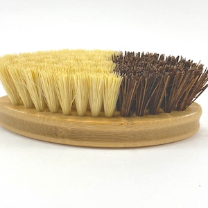 May include: A wooden oval-shaped brush with two sides. One side has soft, light brown bristles and the other side has stiff, dark brown bristles.
