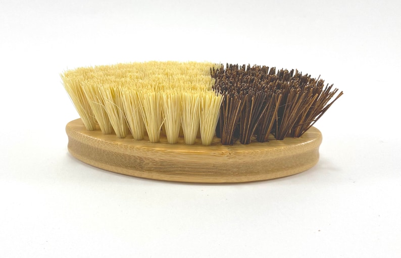 May include: A wooden oval-shaped brush with two sides. One side has soft, light brown bristles and the other side has stiff, dark brown bristles.